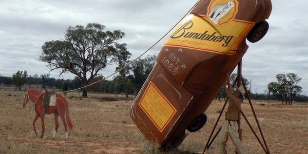 A man in a cowboy hat stands under what seems to be a 12 metre tall, leaning bottle of Bundaberg brand rum, tied to a painted metal horse among scrub and gum trees. The rum bottle is actually a painted sedan-style car. 