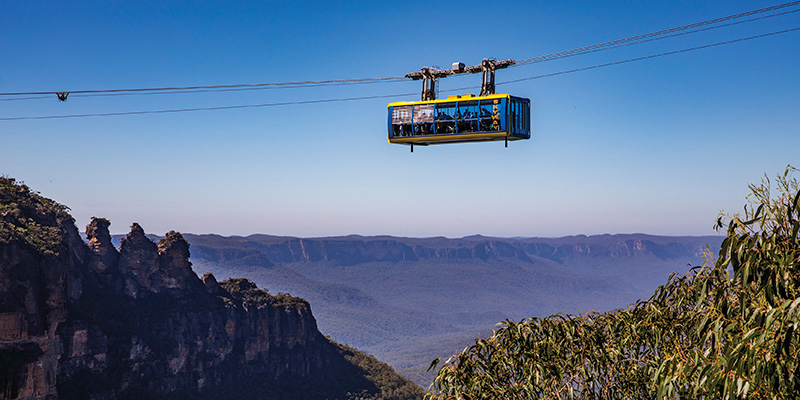 A large yellow passenger cable car with yellow trim and windows on all sides suspended in blue sky above trees and a deep valley.