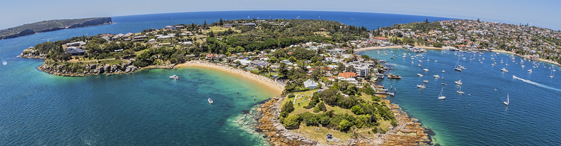 bondi-manly-walk-1920x500 An near-aerial panoramic view of a peninsula of two heads of land with low-lying buildings, leading to two bays with beaches. Boats float just offshore in clear blue water.