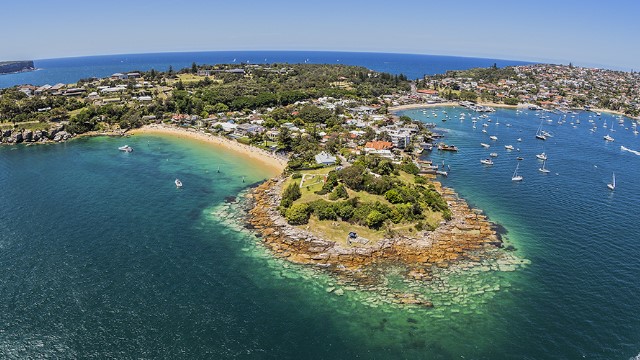 An near-aerial panoramic view of a peninsula of two heads of land with low-lying buildings, leading to two bays with beaches. Boats float just offshore in clear blue water.