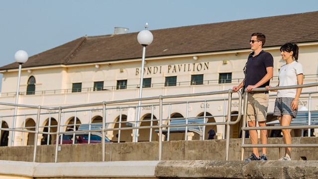 A man and woman lean against a metal railing along a boardwalk lined with vintage, round street lamps, on a cloudy day. Behind them is a long off-white building with the 'Bondi Pavilion' written in upper case New Roman font above the top row of windows.