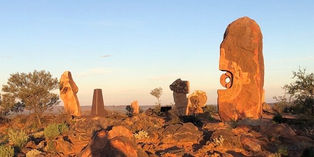 sun shines on an ocre-coloured rock with a round cutout sitting on rocky ground with blue sky and other rock sculptures in the background