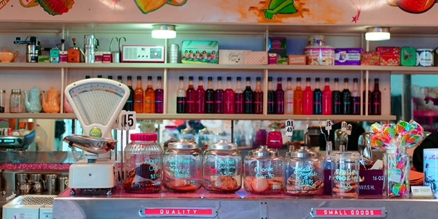 old fashioned counter with scales and jars with biscuits and lollies with bottles of colourful drinks on a shelf behind