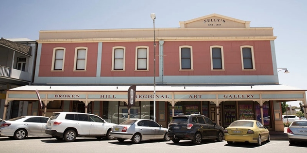 Cars parked at 90 degrees at the front of a Historic two-storey building with pink render and a large awning with signage that says Broken Hill Regional Art Gallery