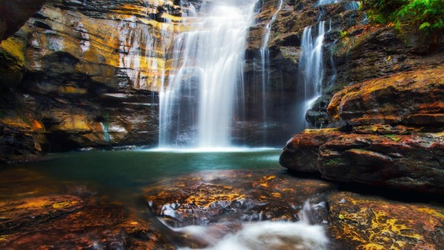 A waterfall lit with sunlight as it pours into a cave over rocks and into a dark pool of green, surrounded by mottled red, brown and yellow cave walls.  