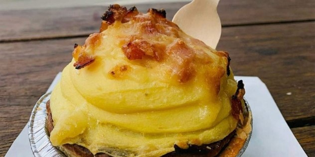 Close up of a meat pie on a wooden outdoor table, the pie is in a metal tin and is topped with a swirl of mashed potato and bacon bits. 