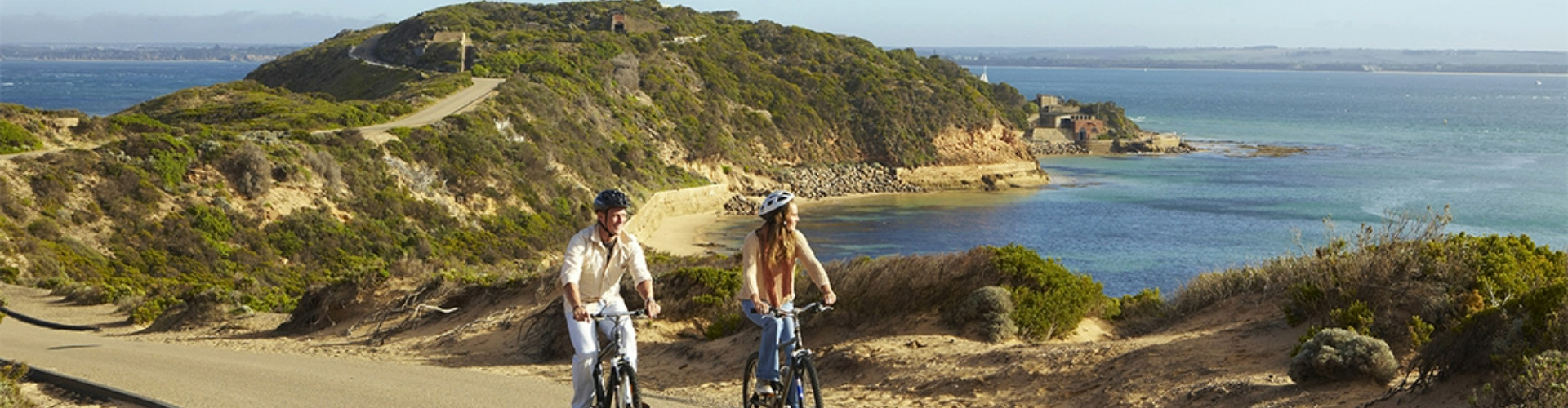 cyclists-at-mornington-peninsula-vic-1920x500 A couple cycling a sunny bike path up the shoreline from a green covered peninsula behind them.