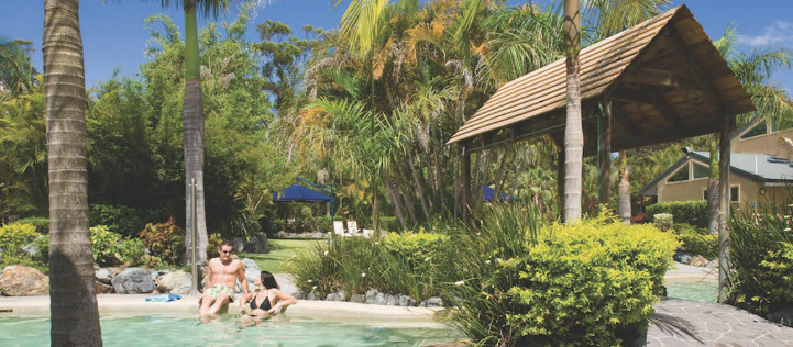 A couple relaxes in a tropical, landscaped resort pool area under palm trees and blue sky.