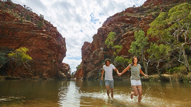 a happy couple run through ankle deep water in a creek with large red rock formations in the background and gum trees on the sides of the creek