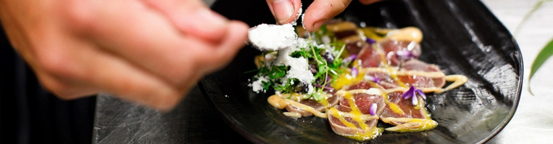 entree-at-hungry-duck-berry-nsw-1920x500 A hand holds a spoon tapping white powder over greens and thin sliced raw tuna drizzled with yellow sauce on a black plate.