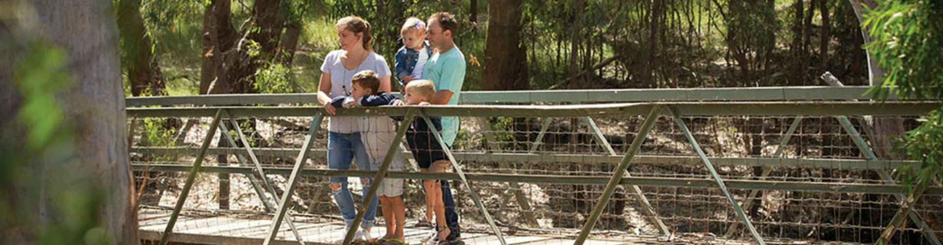 family-at-island-sanctuary-nsw-1920x500 A couple, with a baby and two kids, standing on a walking bridge in a sunlit forest.