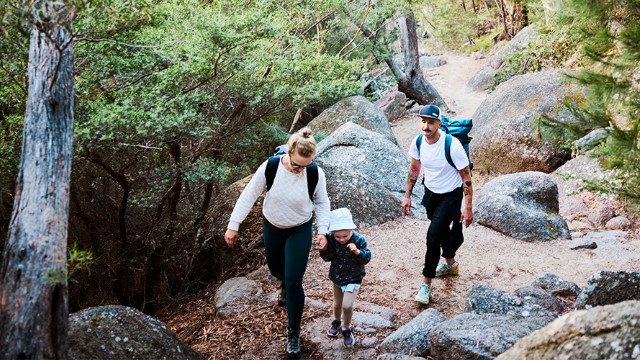 family-bushwalk-hike-640x360 A blond mom in a backpack holds a toddler's hand, followed by a mustached dad, as they walk up a path surrounded by large rocks and trees.