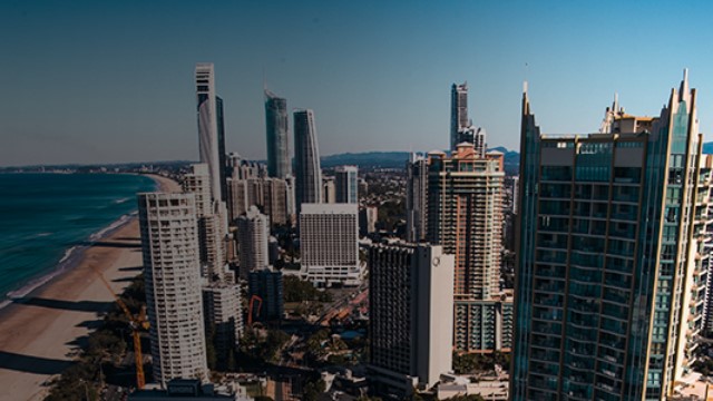 A city with several skyscrapers along a beach, as seen from another skyscraper window, framed by the ocean and mountains on the horizon.
