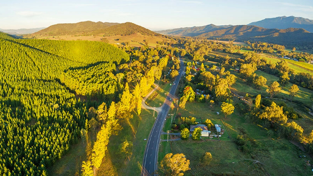 A panoramic view of a road running through tall, sunlit, tree-covered hills leading to a mountain range in the distance. 