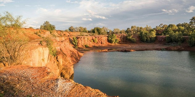 A small, rounded, orange-rock walled gorge around a dark blue pond in the outback, with patches of trees along one side. This is part of the Cobar Heritage Walk.  