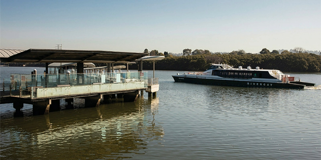 A covered, flat top pier with glass railings stretches out into a mangrove-lined river, as a passenger boat goes by.