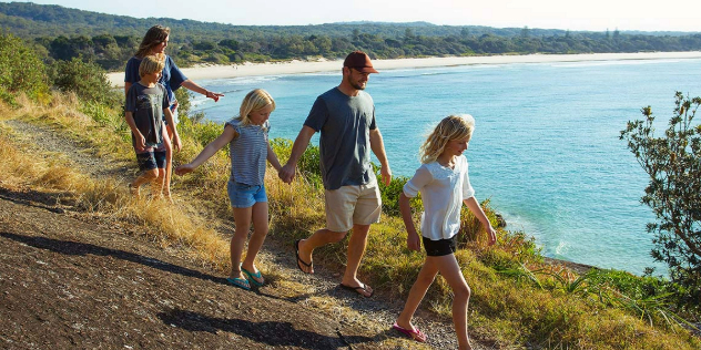  A family walking along a path on a grassy cliff's eduge, over a sunny, white sand beach and forested shores.