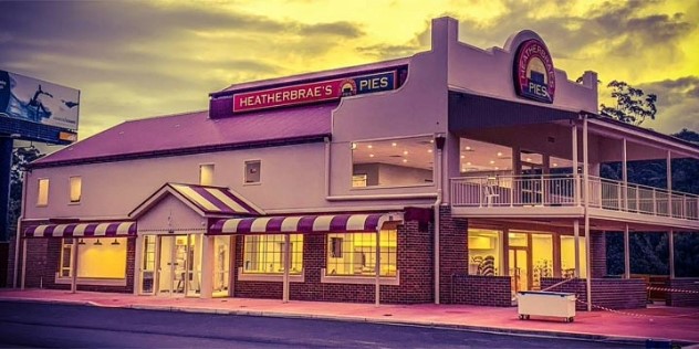 An older two-story pub style building with a striped awning out front, and a Heatherbrae’s Pies sign on the top level, all tinted in purple and yellow sunset light. 