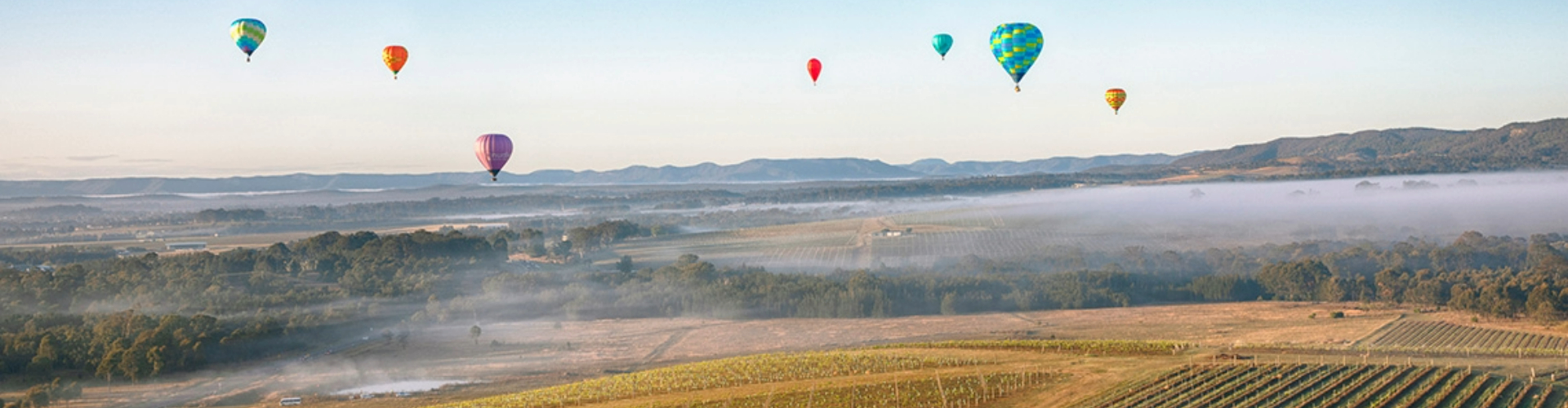 hot-air-balloons-hunter-valley-nsw-1920x500 Seven colourful hot air balloons hover in blue sky over paddocks, trees and plains with mountains on the horizon.