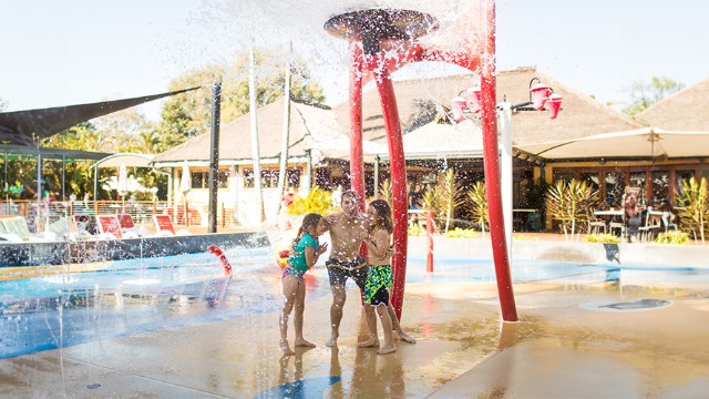Two boys and a girl laugh under a water feature spray at a sunny resort pool.  