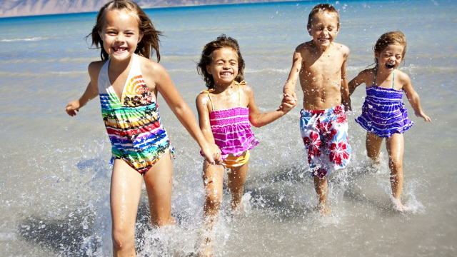 Three girls and a boy in swimsuits laugh and hold hands as they run along a sunny beach.