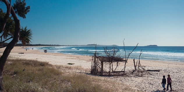 people walking along white sandy beach towards a driftwood construction with the ocean to the right