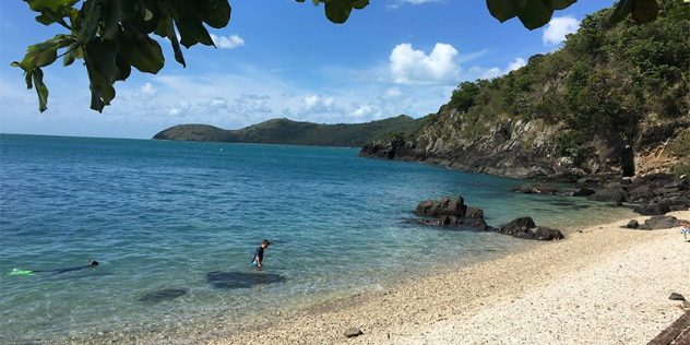  Two people snorkeling along a nearly white sand beach, dotted with rocks and green hilled shores.