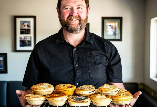  A smiling middle-aged man with brown-red hair and a beard stands in a café holding a metal tray stacked with eight golden baked pies. 