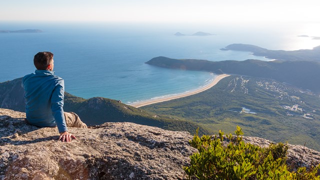 A man sits atop a cliff on a sunny day, leaning back on one hand to overlook a tropical scene of green hills leading to buildings and a beach. 
