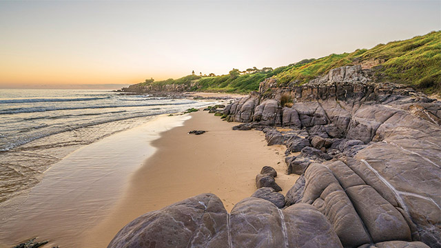 A rocky and sandy beach framed by green shoreline, bathed in a sunset glow. 