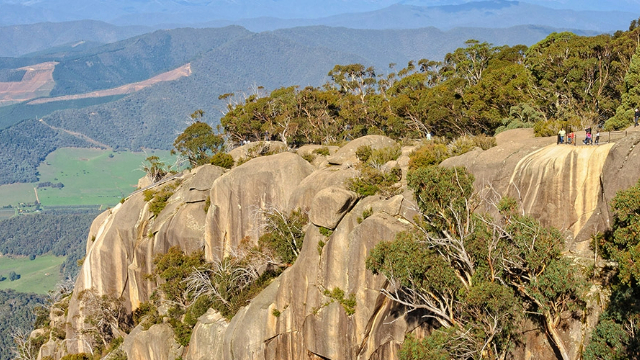 The top of a mountain high above green fields below, covered with rounded rocks and trees.