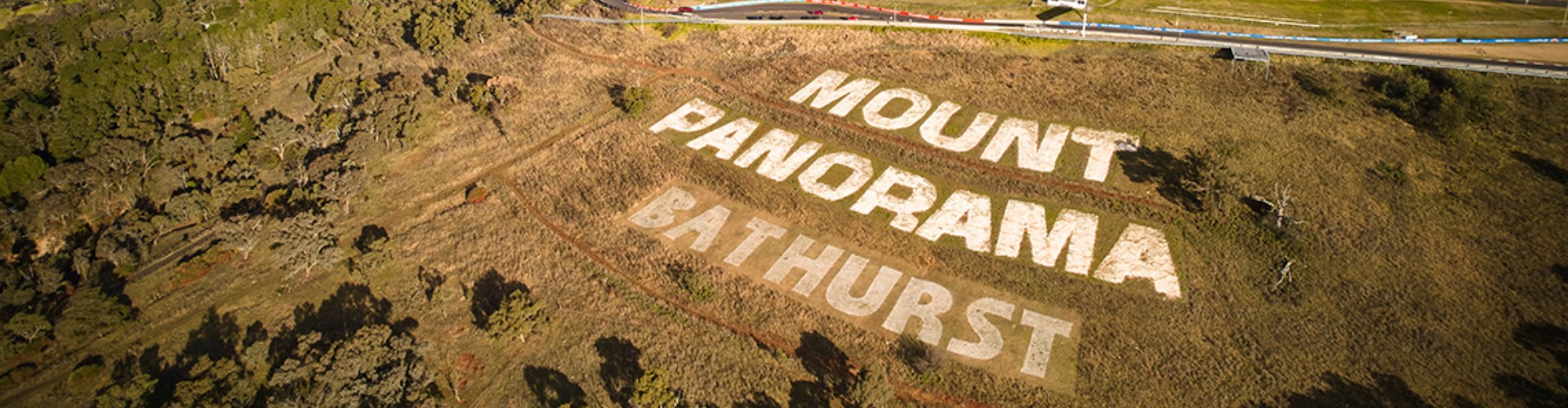 mount-panorama-bathurst-nsw-1920x500 An aerial view of the words Mount Panorama Bathurts mowed into the grass on a sunny hillside next to a road.