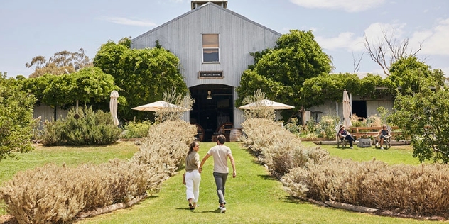 couple holding hands walk down grass pathway flanked with hedges towards door of quaint timber barn