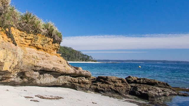  A small white sand beach, hugged by a rocky outcrop of orange rocks, past which a small island covered in trees can be seen in the open ocean, under a blue sky. 
