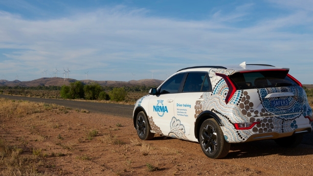 nrma electric vehicle parked at the side of the road facing a long flat road with wind turbines on a hill to the right