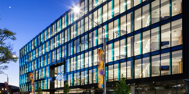 A rectangular office front next to a tree filled with evenly spaced windows, all lit up at dusk.
