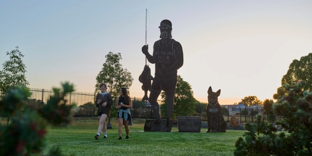 two women chatting as they pass the large steel sculpture of a man wearing a cap and vest holding a fish and rod next to the sculpture of a cattle dog