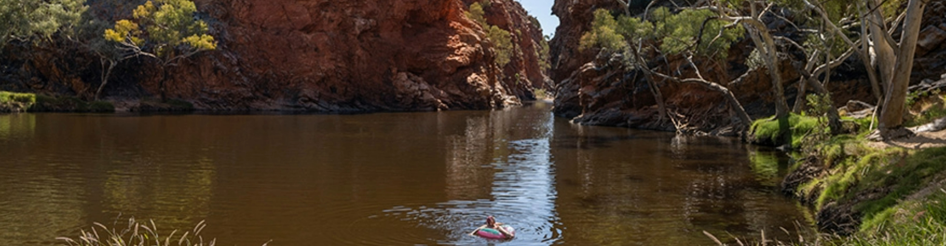ormiston-gorge-waterhole-nt-1920x500 A person floats alone on a tube in a sunny waterhole, surrounded by rocky, red gorge walls and trees onshore.