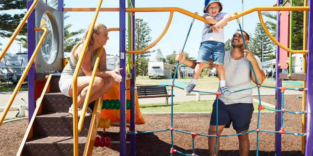 A woman watches a man help a toddler climb a rope ladder on a sunny, colourful playground.