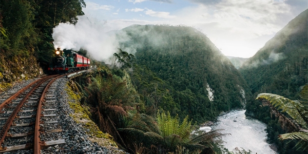 green puffing steam train pulling red carriages rounds the bend next to a tree-covered gorge and river