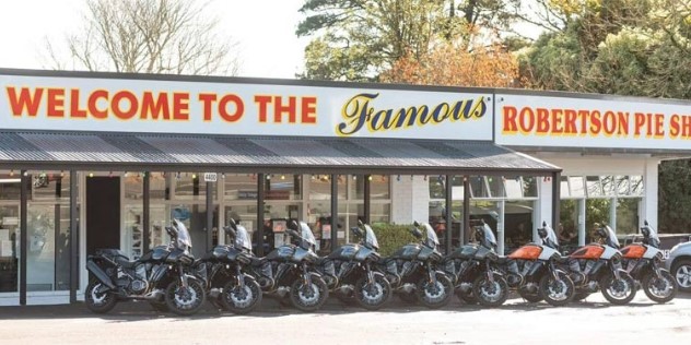 A low-set flat topped café on a sunny day, with Welcome to the Famous Robertson Pie Shop in large font above the doors and a row of nine similar motorcycles parked out front.