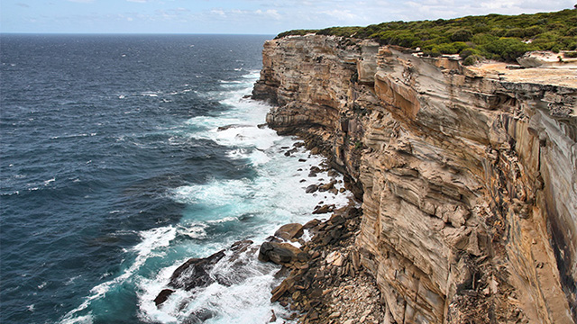 A steep cliffside with horizontally eroded layers of rock, topped with thick grass, stretching into a dark blue sea.