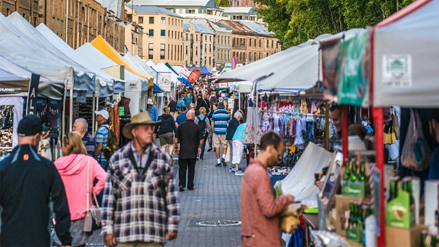 Crowds walk through market tents, with a row of historic buildings up the street behind them.