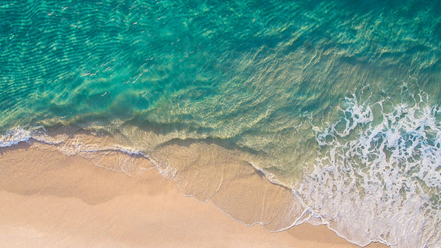 Aerial view of a turquoise blue and foamy wave washing over sand, nothing else.