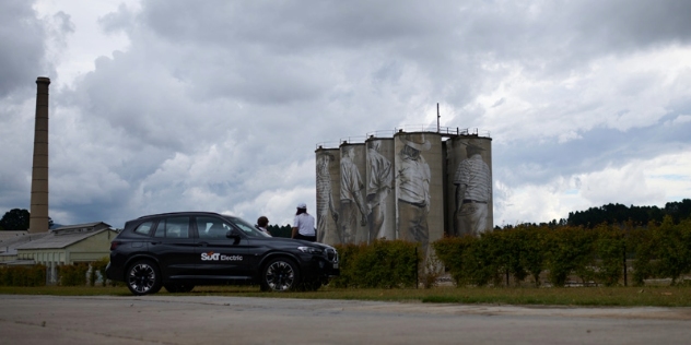 black vehicle with SiXT electric logo on the door parked at the side of the road next to iconic silos painted with images of portland cement workers