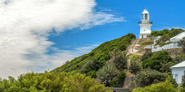 A white lighthouse at the top of a green hill to the right of the frame, the rest filled with clouds and bright blue sky.