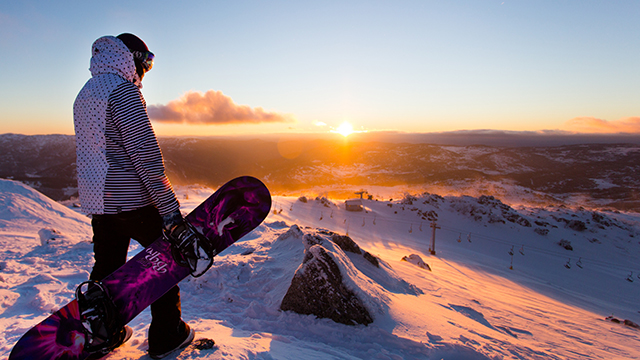 A snowboarder in a hoodie on a mountaintop, looking over peaks bathed in orange sunset light.