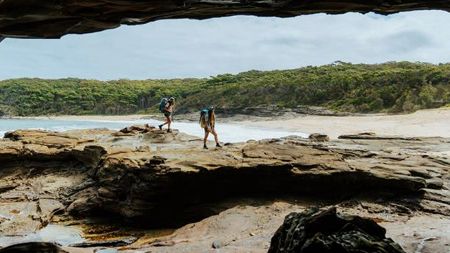 Two hikers traverse flat rock formations just outside caves along a sandy beach with a lush, grassy shore.