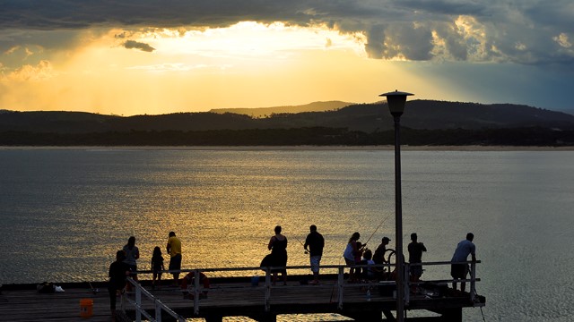 A single bright shaft of yellow sunset light breaks through dark skies, silhouetting rolling hills across the water from Merimbula pier. 