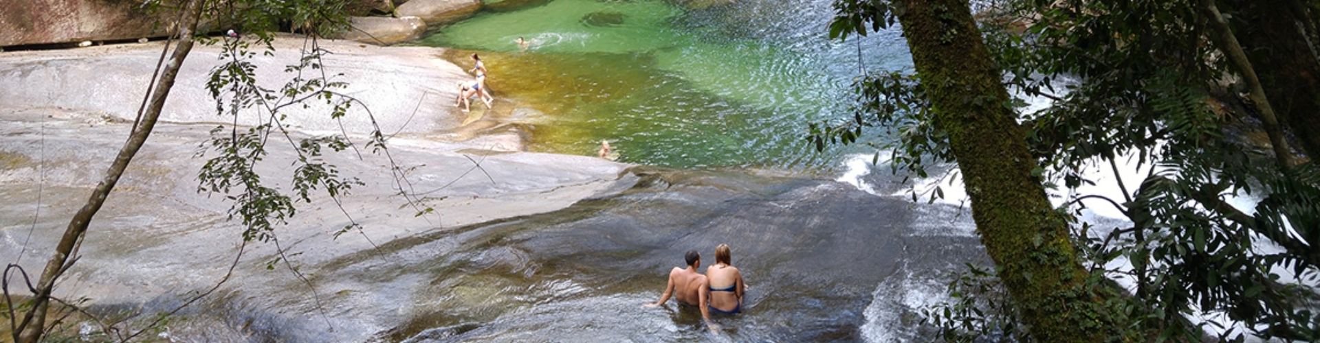 Sustainable travel getty images man and woman in swim suits sit in shallow water on smooth rock at peaceful rocky pool surrounded by rainforest while children swim and play in the water